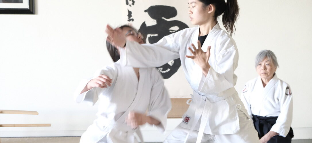 A young aikidoka executes a technique as her partner takes the fall and a senior teacher watches from seiza.