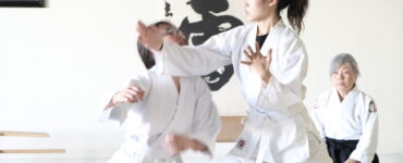 A young aikidoka executes a technique as her partner takes the fall and a senior teacher watches from seiza.