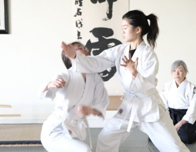 A young aikidoka executes a technique as her partner takes the fall and a senior teacher watches from seiza.