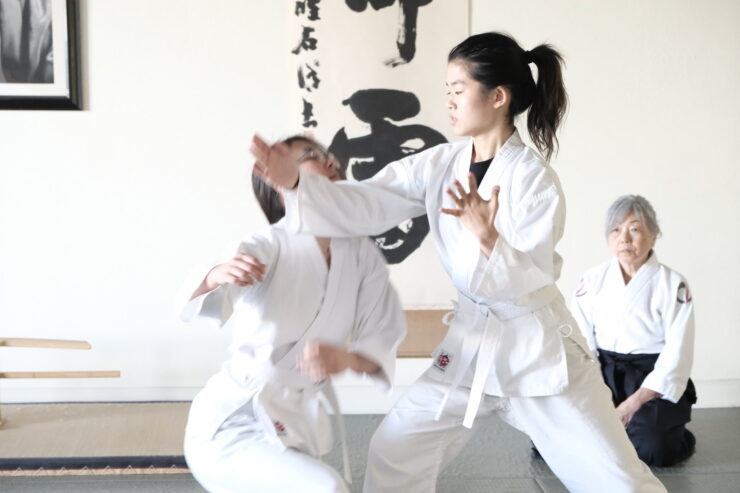 A young aikidoka executes a technique as her partner takes the fall and a senior teacher watches from seiza.