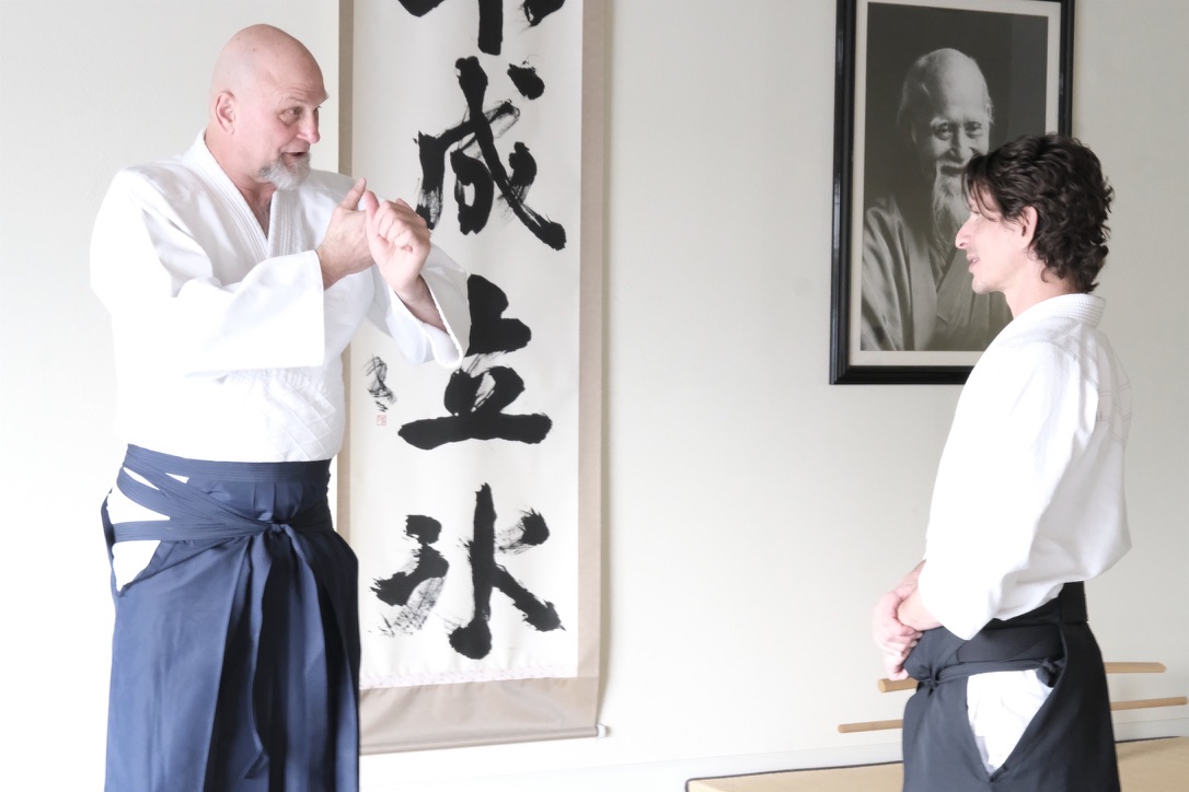 Ellis Amdur, in hakama, explains a point to Josh Gold beneath calligraphy and a portrait of the Founder.